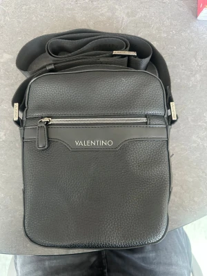 Svart axelväska från Valentino - Snygg svart axelväska äkta från Valentino Mario Valentino i strukturerat konstläder. Väskan har justerbar axelrem, en dragkedjeficka framtill och diskret logga i silver. Perfekt för att bära det viktigaste och ger en stilren look till din outfit. Priset kan diskuteras och kom dm för mer bilder