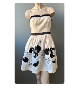 Champagnefärgad kort klänning med svarta detaljer - Snygg kort klänning i champagnefärg med svarta axelband och kant, samt svart band i midjan. Klänningen har blommigt mönster och paljettdetaljer nertill. Tillverkad i glansigt material och har smala axelband.