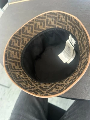 Fendi bucket hat med FF-mönster - Äkta !!!!!!Säljer en brun och svart bucket hat från Fendi med det klassiska FF-logomönstret över hela hatten. Hatten har en mjuk form och är tillverkad i tyg med en beige kant runt brättet. Perfekt för dig som vill ha en statement-accessoar.