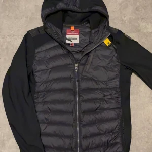 Svart pufferjacka från Parajumpers - Snygg svart pufferjacka från Parajumpers med huva och dragkedja framtill. Jackan har quiltad front, släta ärmar och axlar samt en gul detalj vid kragen. Märkespatch på ärmen och innerfoder med 'WARM UP'-etikett. Perfekt för kalla dagar.