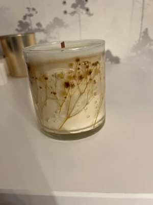 Doftljus i glas med torkade blommor - Unikt doftljus i genomskinligt glas med torkade små blommor inbäddade i vaxet. Ljuset har en träveke och vaxet är vitt med inslag av beige. Perfekt som dekoration eller för mysig stämning hemma.