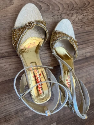 Guldglittriga klackar med pärldetaljer - Eleganta pumps i guld med öppen tå och glittrande pärldetaljer längs remmen. Skorna har en glänsande guldfärgad innersula och smal ankelrem för extra stil. Perfekta för att sticka ut med en lyxig känsla. Klackhöjd cirka 6cm. Köptes utomlands och har använts ett par gånger. Skriv gärna om du har frågor eller vill se ytterligare bilder. Det står storlek 8 på klacken och passar en med storlek 37/38.
