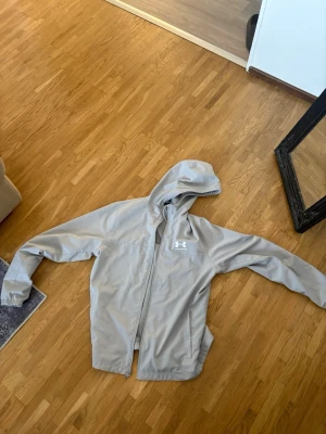 Ljusgrå hoodie från Under Armour - Ljusgrå hoodie från Under Armour med dragkedja och huva. Tröjan har meshfoder på insidan, två sidofickor och elastiska muddar. Perfekt för dig som gillar sportig stil och vill ha något bekvämt och snyggt.