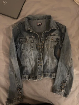Ljusblå jeansjacka med knappar - Snygg ljusblå jeansjacka med klassisk krage och knappstängning framtill. Jackan har två bröstfickor med lock och knappar samt dekorativa sömmar. Perfekt för en avslappnad och trendig look. Den är i strl M men sitter perfekt på mig som har S/xs 