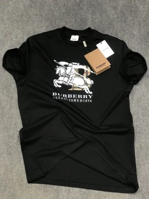 Svart Burberry t-shirt med tryck - Snygg svart t-shirt från Burberry med stort vitt tryck av riddare och logga på bröstet. Klassisk rund hals och korta ärmar. Perfekt för dig som vill ha en stilren och trendig look med exklusiv känsla.