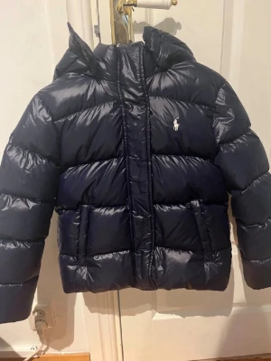 Mörkblå pufferjacka från Ralph Lauren - Snygg mörkblå pufferjacka från Ralph Lauren med huva och klassisk logga på bröstet. Jackan har en glansig finish, breda kanaler och två stora fickor framtill. Perfekt för kalla dagar och riktigt varm tack vare det vadderade materialet.