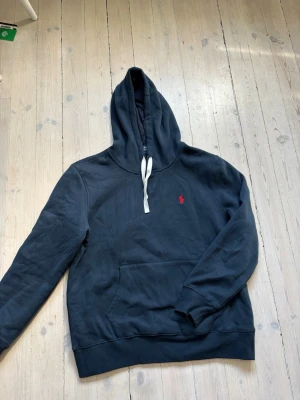 Marinblå hoodie från Polo Ralph Lauren - Snygg marinblå hoodie från Polo Ralph Lauren i storlek M, slim fit. Klassisk design med huva och vita dragsnören samt känguruficka framtill. Ikonisk röd logga broderad på bröstet. Tillverkad i mjuk bomullsblandning för extra komfort.