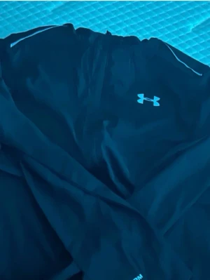Svart vindjacka från Under Armour - Snygg svart vindjacka från Under Armour med reflekterande detaljer på axlarna och stort logotryck på ryggen. Jackan har dragkedja framtill och är tillverkad i lätt, vindtätt material som passar perfekt för aktiva dagar utomhus.