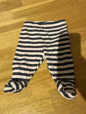 Randiga babybyxor med fot från Cubus - Supersöta babybyxor från Cubus i storlek 50, upp till 2 månader. Byxorna har svarta och ljusrosa ränder samt inbyggda fötter. Endast använda på dockor. Skickar helst med PostNord😄