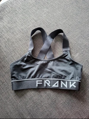 Svart sport-bh från Frank Dandy - Snygg svart sport-bh från Frank Dandy med bred resår och logga framtill och baktill. Bh:n har korsade axelband och är tillverkad i ett stretchigt, ventilerande material som passar perfekt för träning eller en aktiv vardag.