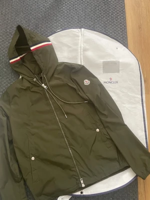 Olivgrön vindjacka från Moncler - Snygg olivgrön vindjacka från Moncler med huva och dragsko. Jackan har hel dragkedja, två fickor med tryckknappar och Moncler-logga på ärmen. Materialet är tunt och vattenavvisande, perfekt för blåsiga dagar. Röd, vit och blå detalj på huvan. Skriv till om ni har några funderingar!