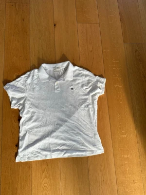 Vit pikétröja från Gant - Klassisk vit pikétröja från Gant med broderad logga på bröstet. Tröjan har krage, korta ärmar och knappar vid halsen. Perfekt till jeans eller shorts för en clean och stilren look.