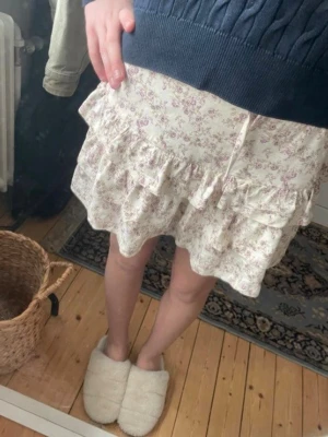 Blommig volangkjol i vitt och lila - Supersöt kort kjol med blommigt mönster i vitt och lila. Kjolen har flera volanger och en flowy passform som ger en romantisk vibe. Perfekt att matcha med en stickad tröja eller en enkel topp. Ungifär 152 i storlek. Vinns en liten gul fläck 