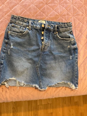 Blå jeanskjol från ONLY med knappar - Säljer en blå jeanskjol från ONLY med rå kant nertill och slitna detaljer. Kjolen har framfickor, bakfickor och synliga guldknappar framtill. Perfekt för en avslappnad och trendig look.