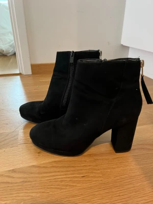 Svarta stövletter med klack - Säljer ett par svarta stövletter i mockaimitation med chunky klack och dragkedja både på sidan och bak. Snygg, stilren design som passar till många outfits. Perfekta för dig som gillar en edgy look och vill ha lite extra höjd. Endast använda en gång, som nya!
