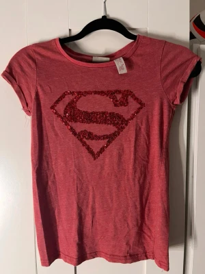 Röd Superman t-shirt med paljetter - Cool röd t-shirt med glittrande Superman-logga i paljetter på bröstet. T-shirten har rund halsringning och korta ärmar. Perfekt för dig som vill sticka ut med en superhjälte-look. Materialet känns mjukt och skönt mot huden. Storleken är xs/158