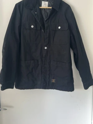 Svart jacka från H&M med fickor - Svart jacka från H&M med fyra stora fickor framtill och silverfärgade knappar. Jackan har klassisk krage och är tillverkad i ett kraftigt bomullsmaterial. Perfekt för dig som vill ha en enkel och stilren jacka med praktiska detaljer.