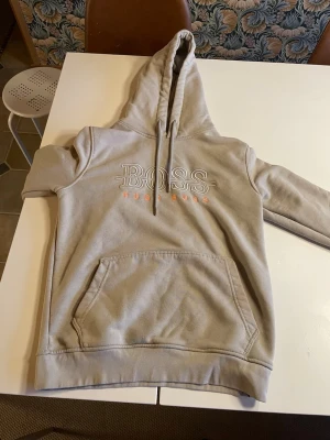 Beige hoodie från Hugo Boss - Snygg beige hoodie från Hugo Boss med broderad logga i vitt och orange på bröstet. Hoodien har en stor huva med dragsko och en praktisk magficka. Perfekt för dig som gillar stilrena och exklusiva plagg.