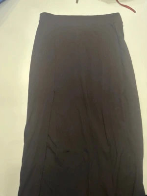 Svart lång kjol i stretchigt tyg med öppet ben - En svart lång kjol med rak passform och öppet ben på ena sidan. Kjolen är gjord i ett stretchigt material som ger en bekväm känsla och följer kroppen snyggt. Perfekt att styla med sneakers eller boots för en cool look. Storlek M från bikbok🥰