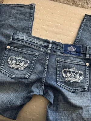 Blåa jeans med kronbrodyr från Victoria Beckham  - Snygga blå jeans från Victoria Beckham med slitningseffekt och vita broderade kronor på bakfickorna. Klassisk femficksmodell med låg midja och raka ben. Perfekta för dig som vill sticka ut med coola detaljer. Bootcut och Lågmidjade.  Midja: 38cm Innerben: 80cm Gren: 17cm