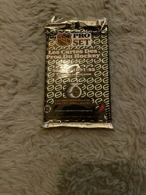 NHL Pro Set hockeykort 1991-92 - Oöppnat paket med NHL Pro Set hockeykort från säsongen 1991-92. Förpackningen är svart och silver med officiell NHL-logga och innehåller 15 samlarkort på franska med statistik och bilder. Perfekt för dig som gillar samlarkort och hockey.