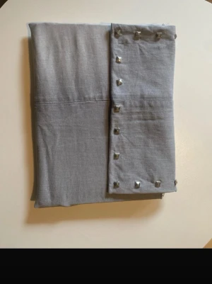 Ljusgrå datorfodral med nitar - Säljer en ljusgrå datorväska i mjukt tyg med silverfärgade nitar längs ena kanten. Väskan har en enkel, rektangulär form och ger en cool touch till din outfit. Perfekt att hålla för en trendig look.