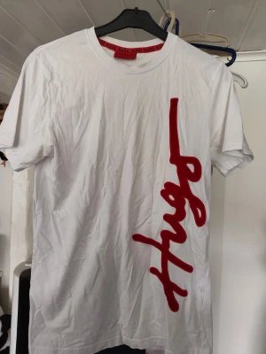 Vit t-shirt från Hugo med röd logga - Vit t-shirt från Hugo med stor röd logga i sammetsliknande material längs ena sidan framtill. Klassisk passform med korta ärmar och rund halsringning. Perfekt för en trendig och avslappnad stil.