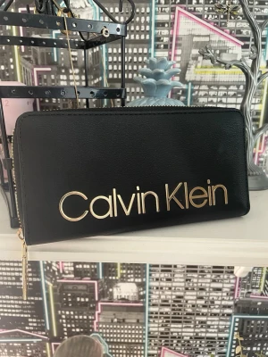 Äkta svart plånbok från Calvin Klein - Snygg svart plånbok från Calvin Klein i slätt konstläder med stort guldfärgat logotyptryck på framsidan. Plånboken har dragkedja med gulddetaljer och Calvin Klein-gravyr på zip-pullern. Perfekt för att hålla koll på kort och cash. Imprinsip ny 💕