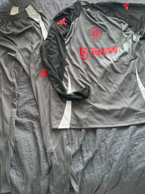 Manchester United träningsset Adidas - Säljer ett grått Manchester United träningsset från Adidas med långärmad tröja och matchande byxor. Setet har svarta och vita detaljer, röda loggor och är gjort i ett lätt, snabbtorkande material. Perfekt för fotbollsträning eller gymmet.