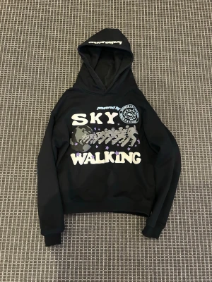Svart hoodie Broken Planet Sky Walking - Svart hoodie från Broken Planet med trycket 'SKY WALKING' i vitt och lila på framsidan. Stor grafisk design med figurer, jordglob och stjärnor. Huva med textdetalj. Perfekt för dig som gillar streetwear och unika prints.