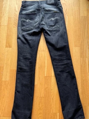 Vintage Nudie Jeans - Snygga mörkgra jeans frän Nudie Jeans i modellen Black Lotus. Slim fit med klassisk femficksdesign som ger en modern, stilren silhuett. Naturliga slitningar ger varje par en unik vintagekansla, medan diskreta kontrastsömmar på bakfickorna forstarker den ikoniska Nudie-stilen