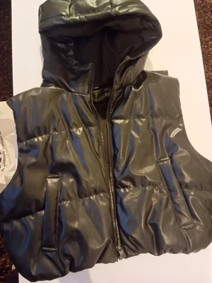 Svart vadderad väst med huva - Snygg svart vadderad väst i glansigt material med dragkedja framtill och två fickor. Västen har en stor huva och en modern, puffig look. Perfekt för lager-på-lager och streetstyle.