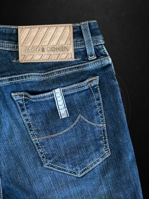 Jacob Cohen jeans  - Snygga mörkblå jeans från Jacob Cohen med klassisk femficksdesign och diskreta slitningar framtill. Jeansen har en rak passform och tydliga kontrastsömmar. Tillverkade i mjuk denim med en beige patch bak i midjan.