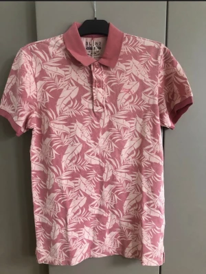 Rosa pikétröja med bladprint från Blend - Snygg rosa pikétröja från Blend med vitblommigt bladmönster. Tröjan har klassisk krage, knappar framtill och korta ärmar. Perfekt för en avslappnad och trendig look.