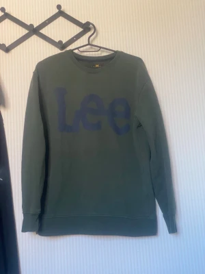 Grön sweatshirt från Lee - Mörkgrön sweatshirt från Lee med stort blått tryck på bröstet. Tröjan har rund halsringning, långa ärmar och ribbade muddar vid ärmslut och nederkant. Perfekt för en avslappnad och cool stil.