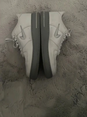 Nike Air Force 1  - Säljer ett par klassiska Nike Air Force 1 sneakers i vitt med grå sula och silverfärgad swoosh. Skorna har snörning och är tillverkade i skinn, vilket ger en clean och tidlös look. Perfekta för dig som gillar streetwear och vill ha något stilrent.