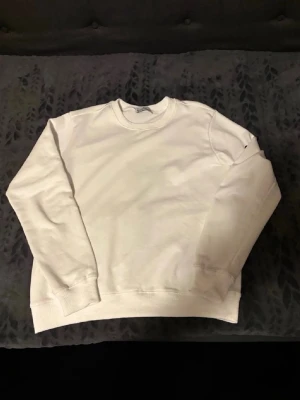 Vit sweatshirt från Stone Island - Säljer en stilren vit sweatshirt från Stone Island. Tröjan har rund halsringning, ribbade muddar vid ärmslut och nederkant samt klassisk passform. Perfekt för dig som gillar enkel och clean stil. Materialet är mjukt och bekvämt bomullstyg.