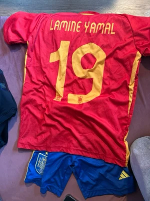 Spanien fotbollströja Lamine Yamal - Röd och gul Spanien fotbollströja från Adidas med Lamine Yamal och nummer 19 på ryggen. Tröjan har gula detaljer och är tillverkad i lätt och ventilerande AEROREADY-material. Perfekt för dig som älskar fotboll och vill stötta Spanien.