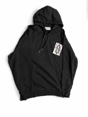 Svart hoodie med spelkortstryck från Saint Laurent  - Cool svart hoodie från Saint Laurent med huva och dragsko. På bröstet finns ett unikt tryck av ett spelkort med motiv i svart, vitt och rött. Hoodien har en stor magficka och ribbade muddar. Perfekt för dig som gillar streetwear och snygga detaljer. Pris 1500 men går att diskuteras vid snaps affär 