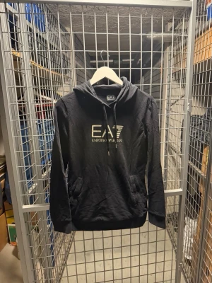 Svart hoodie från EA7 Emporio Armani - Snygg svart hoodie från EA7 Emporio Armani med stor logga i vitt framtill. Klassisk huva med dragsko och känguruficka. Perfekt för en avslappnad och trendig stil. Mjukt material och ribbade muddar vid ärmslut och nederkant.