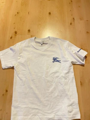Vit Burberry t-shirt med blå logga - Snygg vit t-shirt från Burberry med blått tryck av riddare på bröstet och Burberry-text på ärmen. Klassisk passform och rund halsringning. Tillverkad i mjuk bomull som känns skön mot huden. Perfekt för dig som gillar stilrena plagg med exklusiv touch.
