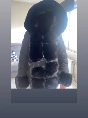 Svart pälspuffjacka med stor huva - Snygg svart pälspuffjacka med stor fluffig huva och breda pälspaneler framtill. Perfekt för kalla dagar och ger en lyxig känsla. Jackan är i en längre modell och har en modern, trendig look. Aldrig någonsin använt!! Lokaliserad i Göteborg