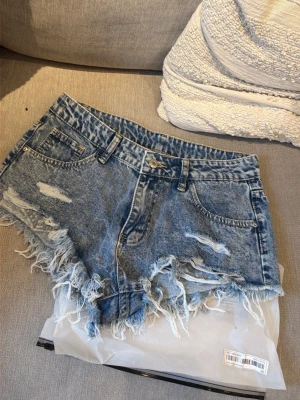 Nya i förpackning! Blå slitna jeansshorts med fransar - Säljer ett par blå denimshorts med slitna detaljer och fransig kant. Klassisk femficksmodell med bälteshällor och knappstängning. Perfekta för en avslappnad och trendig look.