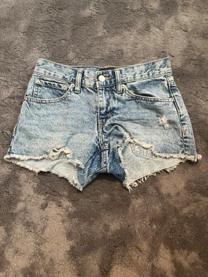 Blå denimshorts med slitningar - Snygga blå denimshorts med råa kanter och slitna detaljer framtill. Klassisk femficksmodell med knapp och dragkedja. Perfekta för sommaren och ger en avslappnad streetstyle-look.