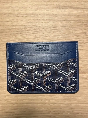 Blå korthållare i skinn/läder - Snygg korthållare i blått skinn med klassiskt mönster i vitt och brunt. Perfekt för kort och ID, med flera fack och stilren design. Märket är präglat på framsidan och den har en lyxig känsla som sticker ut.