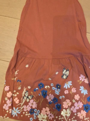 Rostfärgad klänning med blommor - Söt rostfärgad klänning med blommigt mönster och fjärilar nertill. Klänningen är ärmlös och har en avslappnad passform, perfekt för varma dagar. Tillverkad i mjuk bomull som känns skön mot huden.