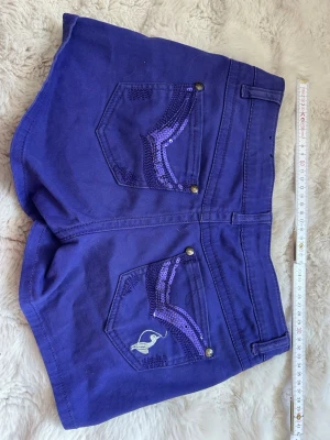 Lila shorts med paljettdetaljer - Snygga lila shorts från Michele Bohbot med paljettdetaljer i olika lila nyanser på fickorna både fram och bak. Klassisk femficksmodell med knapp och dragkedja. Perfekta för dig som vill sticka ut lite extra i sommar!