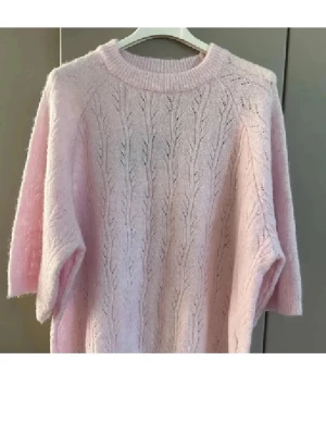 Ljusrosa stickad tröja med mönster - Superfin ljusrosa stickad tröja med kortare ärmar och diskret mönsterstickning. Tröjan har rund halsringning och är gjord i ett mjukt, fluffigt material som känns riktigt mysigt. Perfekt för lager-på-lager och en söt touch till din outfit.