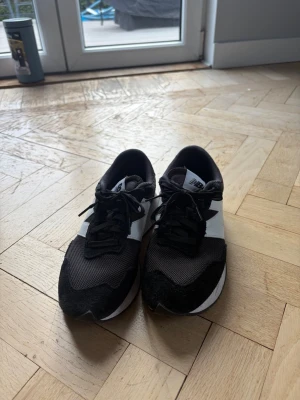 Svarta och vita New Balance sneakers - Snygga New Balance sneakers i svart och vitt med klassisk logga på sidan. Ovandelen är i mesh och mocka för en sportig look och bra andningsförmåga. Skorna har låga skaft och snörning framtill. Perfekta för dig som gillar streetwear och stilren design.