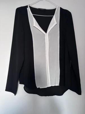Svart och vit blus från Vila - Stilren svart blus från Vila med långa ärmar och vit panel framtill. Djup V-ringning med vit kant ger en snygg kontrast. Tunn och luftig modell som faller snyggt. Perfekt för dig som gillar en klassisk men modern look.